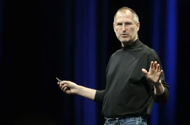 Steve Jobs in a black turtleneck and jeans is speaking on stage, holding a small remote in one hand and gesturing with the other, against a dark background with blue lighting.