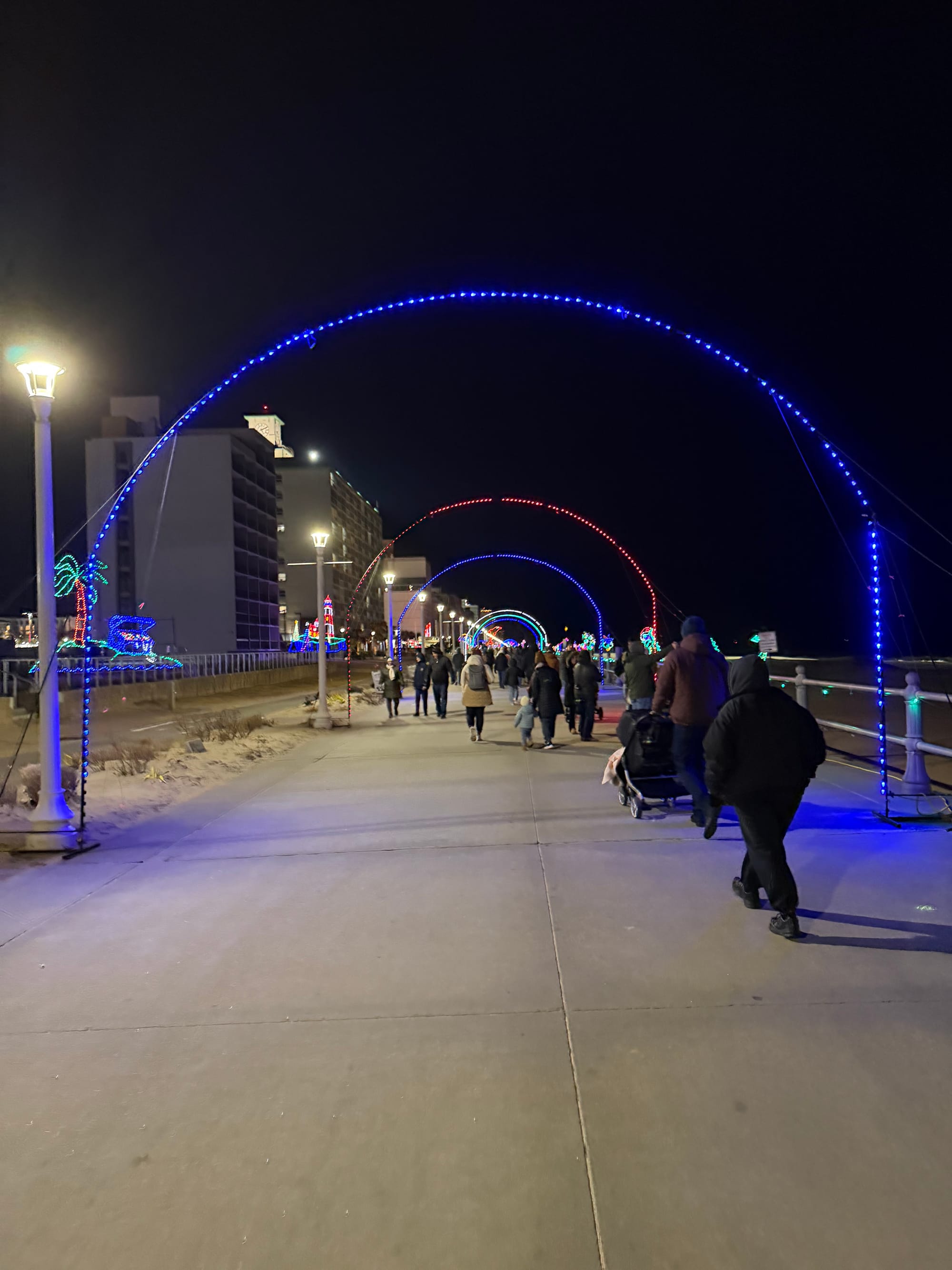 A tunnel of Christmas lights on the boardwalk with hotels on the left and beach on the right.