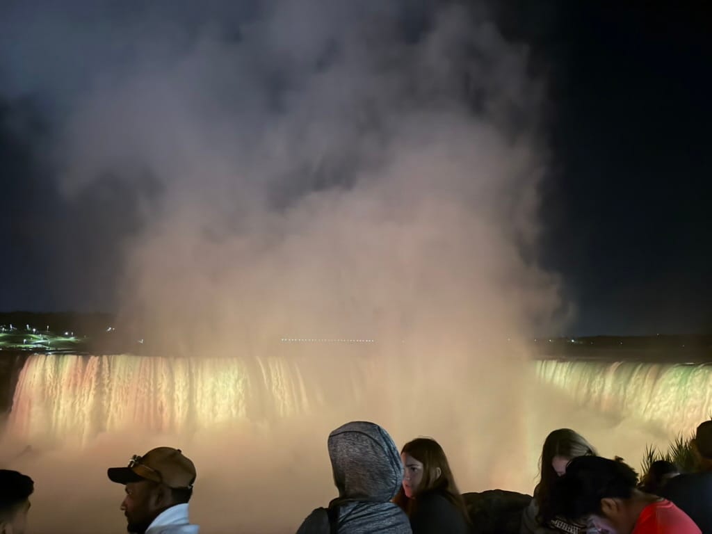 The falls with mist in the front and orange light shining on them in the back.
