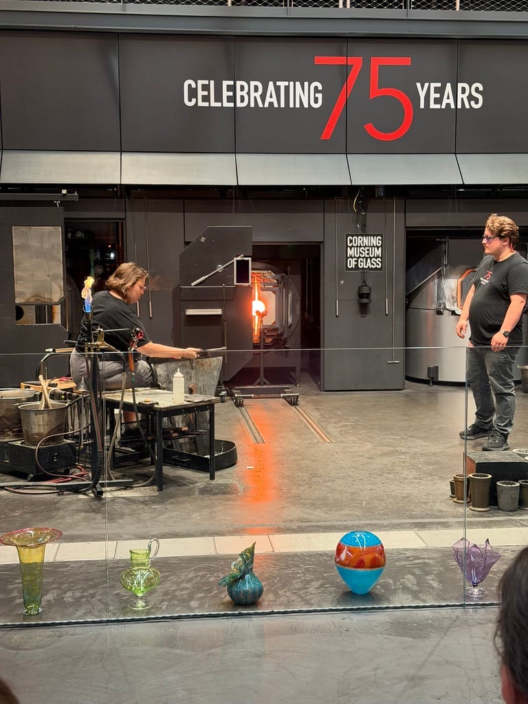 A woman sits, blowing glass, on the left with a man standing at the ready to help on the right.