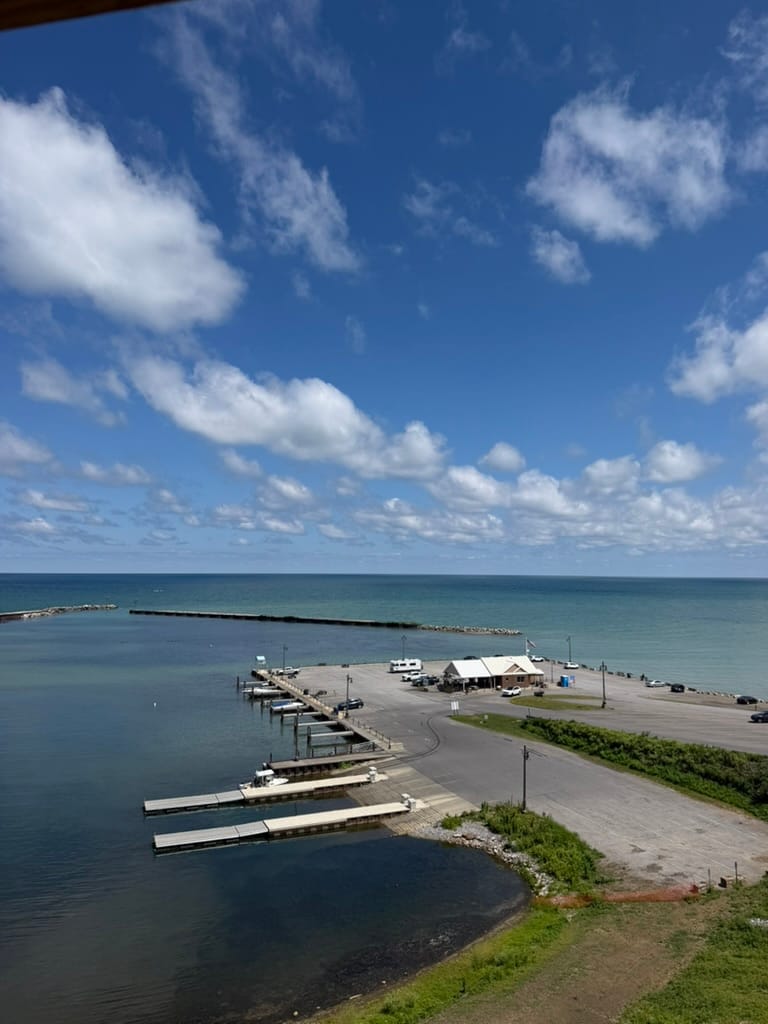 View of the boat landing from the top of the lighthouse. There is a big plot of land with water all around it and building in the middle.