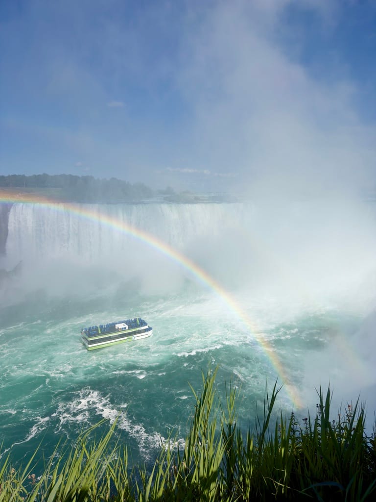 A boat in the center of the falls with a rainbow over the top of it.