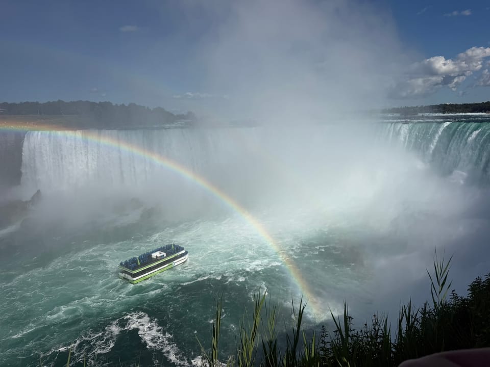 A Niagara boat with in the center of Horseshoe Falls with a rainbow overtop of it. The mist is boiling up from the bottom of the falls.